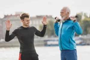 Persona de unos sesenta años entrenando con mancuernas junto a un entrenador al aire libre