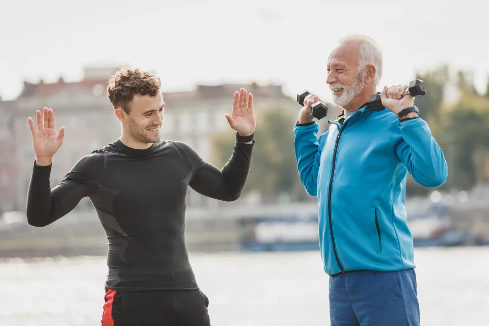 Persona de unos sesenta años entrenando con mancuernas junto a un entrenador al aire libre