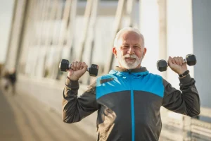 Hombre de unos sesenta años levantando dos mancuernas a la altura de los hombros al aire libre
